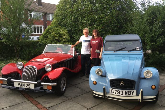 A man and a woman standing next to two classic cars, one is red and one is blue