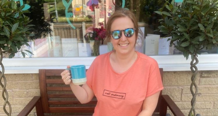 Louise sits on a bench in her garden, holding up a cup of tea to say cheers