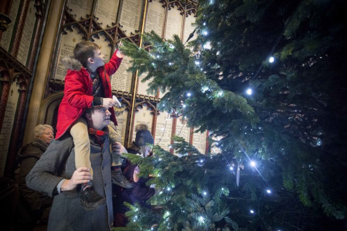 A child sits on their parent's shoulders to put a light on a tall christmas tree in the Cathedral.