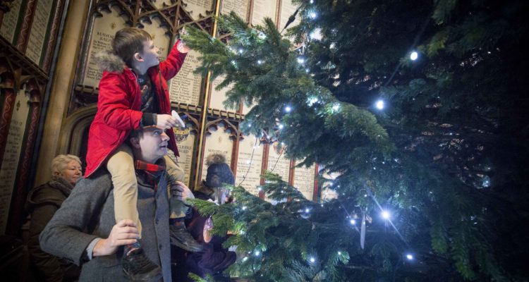 A child sits on their parent's shoulders to put a light on a tall christmas tree in the Cathedral.