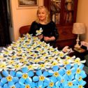 Artist Lindi Kirwin stand behind a giant 3D triangular cake slice, covered with blue metal forget-me-not flowers.