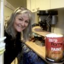 Artist Lindi holding a painted wooden frame around her head, smiling and looking at the camera