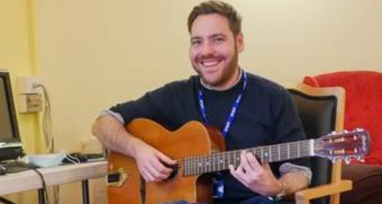 Music facilitator Andy sat on a chair holding a guitar and smiling
